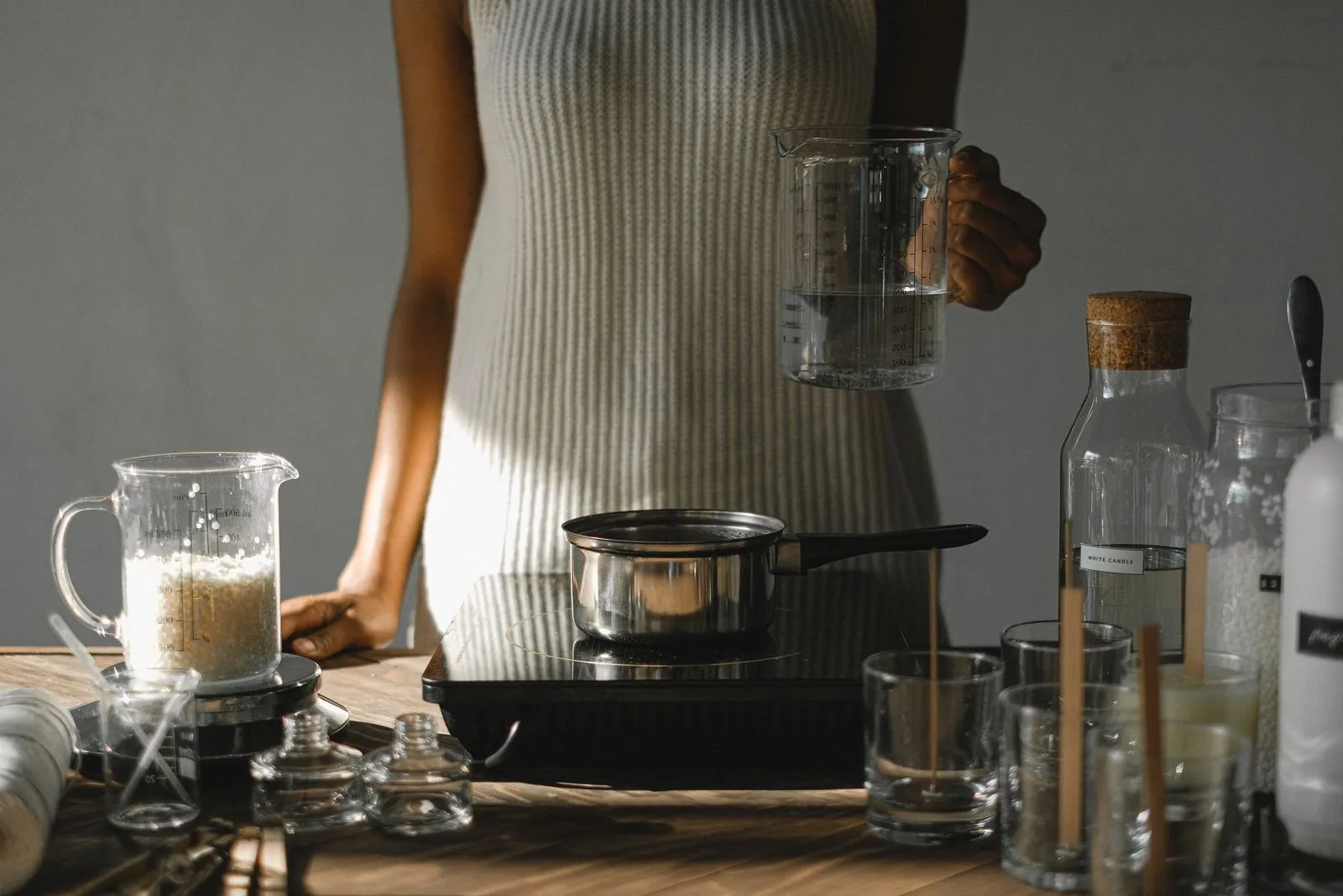 Unrecognizable African American female standing at wooden table with pot on cooker while preparing for melting wax flakes in workshop
